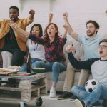 Tips nonton bareng : Sekelompok anak muda sedang bersorak gembira menonton pertandingan bola di depan layar televisi ruang tamu