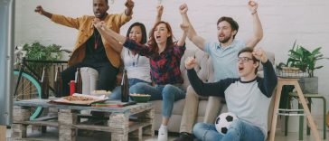 Tips nonton bareng : Sekelompok anak muda sedang bersorak gembira menonton pertandingan bola di depan layar televisi ruang tamu