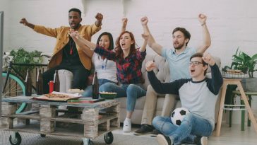Tips nonton bareng : Sekelompok anak muda sedang bersorak gembira menonton pertandingan bola di depan layar televisi ruang tamu