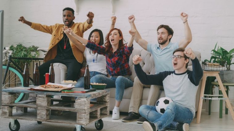 Tips nonton bareng : Sekelompok anak muda sedang bersorak gembira menonton pertandingan bola di depan layar televisi ruang tamu