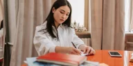 Seseorang mencatat poin evaluasi kerja mingguan di buku catatan meja kantor