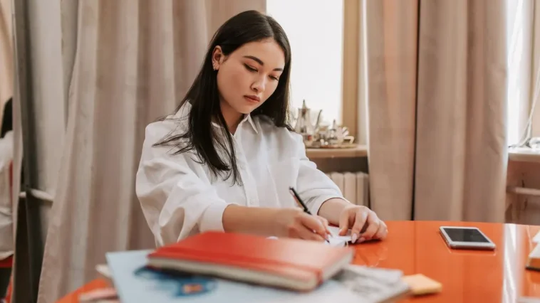 Seseorang mencatat poin evaluasi kerja mingguan di buku catatan meja kantor